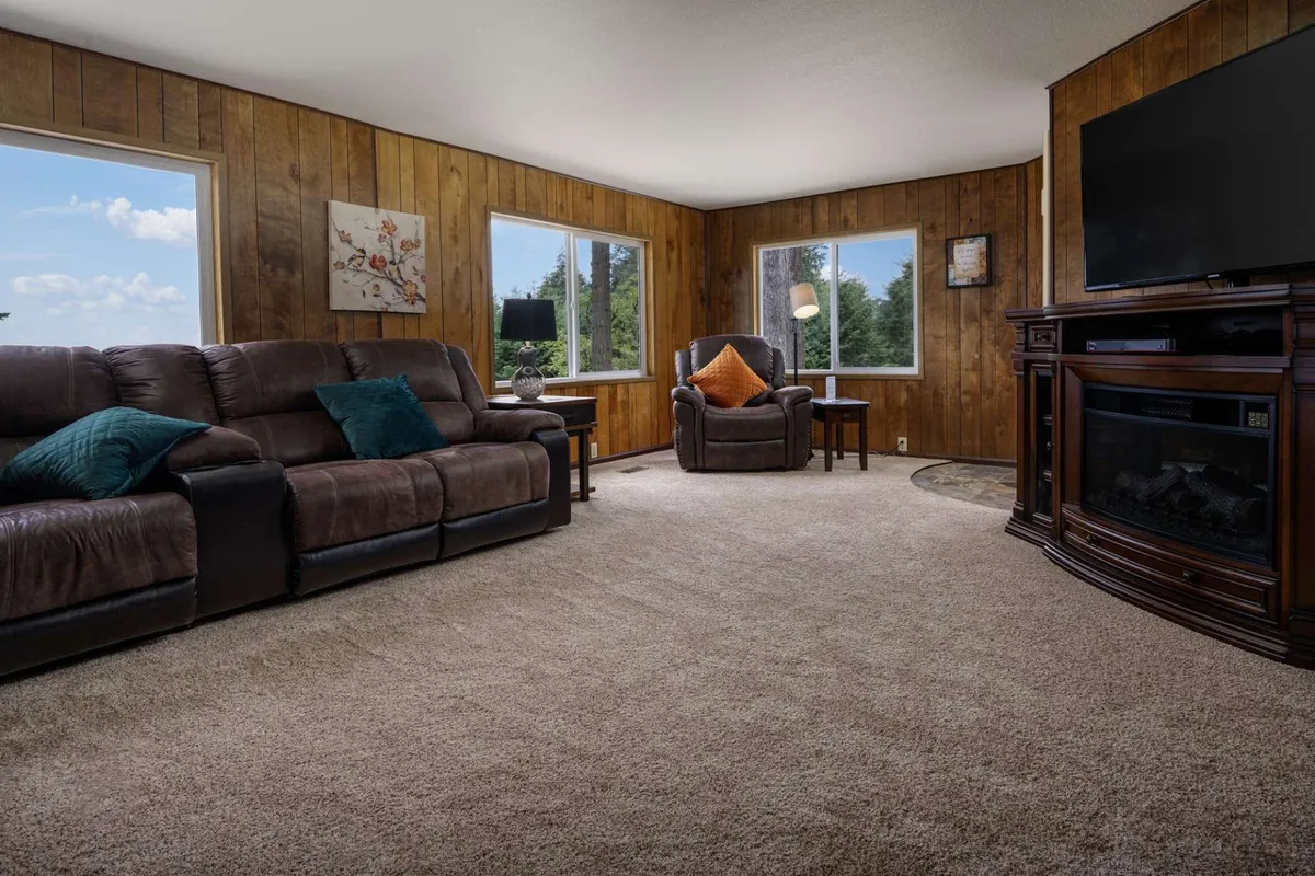 A cozy, sunlit living area featuring a comfortable leather sofa and an armchair with an orange accent pillow. Large windows fill the room with natural light, while a flat screen TV provides entertainment. The relaxed layout encourages socializing and quiet relaxation alike.