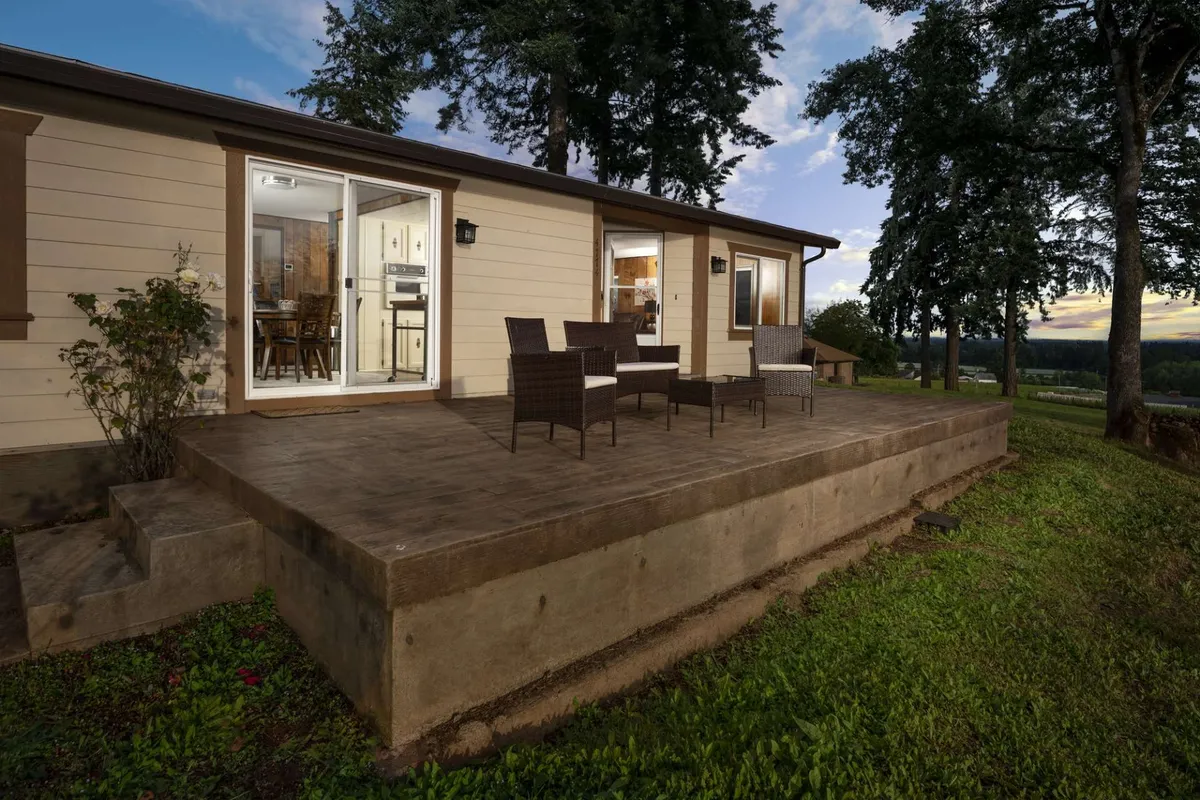 A spacious wooden deck bordered by mature trees, furnished with rattan chairs and a large dining table. The setup provides an inviting space for outdoor dining or lounging, and the deck overlooks expansive landscape views, hinting at vineyard vistas. The environment feels serene, ideal for gathering, sharing meals, or simply relaxing to take in the scenery.