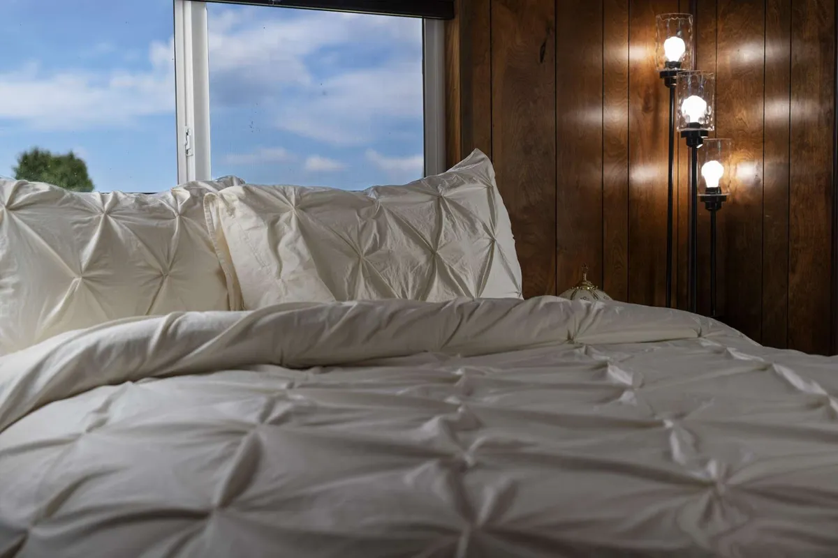 A plush bed with crisp white linens sits beneath a window, inviting in abundant natural light. The view from the window brings the outside in, with unique lighting fixtures adding to the serene atmosphere.