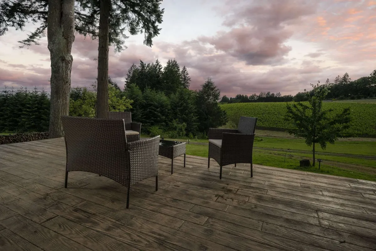 The image showcases the inviting wooden deck of The Blanchard Guest Home, furnished with cozy wicker chairs perfectly positioned to take in sweeping vineyard views. The landscape stretches towards a line of trees under a sky dotted with soft clouds, creating a tranquil atmosphere ideal for relaxation and unwinding with a glass of local wine.