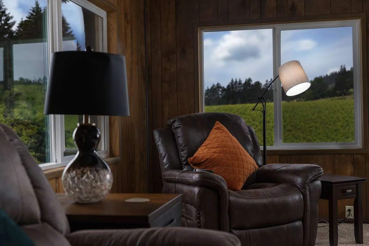 A cozy leather recliner set beside a table lamp, framed by large windows that flood the room with natural light. The windows offer glimpses of lush greenery and the outdoors, suggesting a strong connection to the surrounding landscape.
