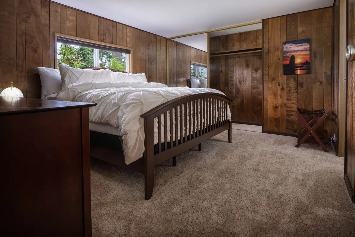 This image features a serene bedroom with a king-size bed dressed in crisp white bedding, flanked by wood paneling and artwork. Ample natural light streams in through large windows, creating a peaceful atmosphere perfect for resting and rejuvenating after a day out.